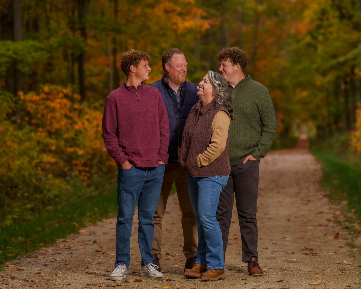 Family of four smiling together on a scenic autumn forest path surrounded by colorful fall foliage.