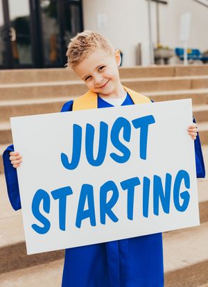kindergarten graduate boy blue cap and gown holding sign "just starting"