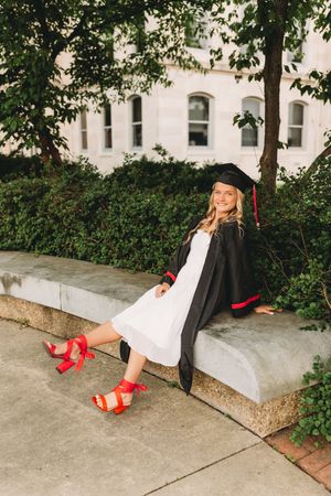 senior graduate girl blonde hair white dress black cap and gown red tassel sitting on bench