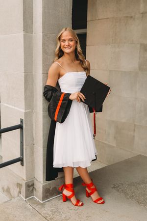 `senior graduate girl blonde hair white dress black cap and gown red tassel stone wall