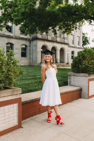 senior graduate girl blonde hair white dress black cap and gown red tassel courthouse building
