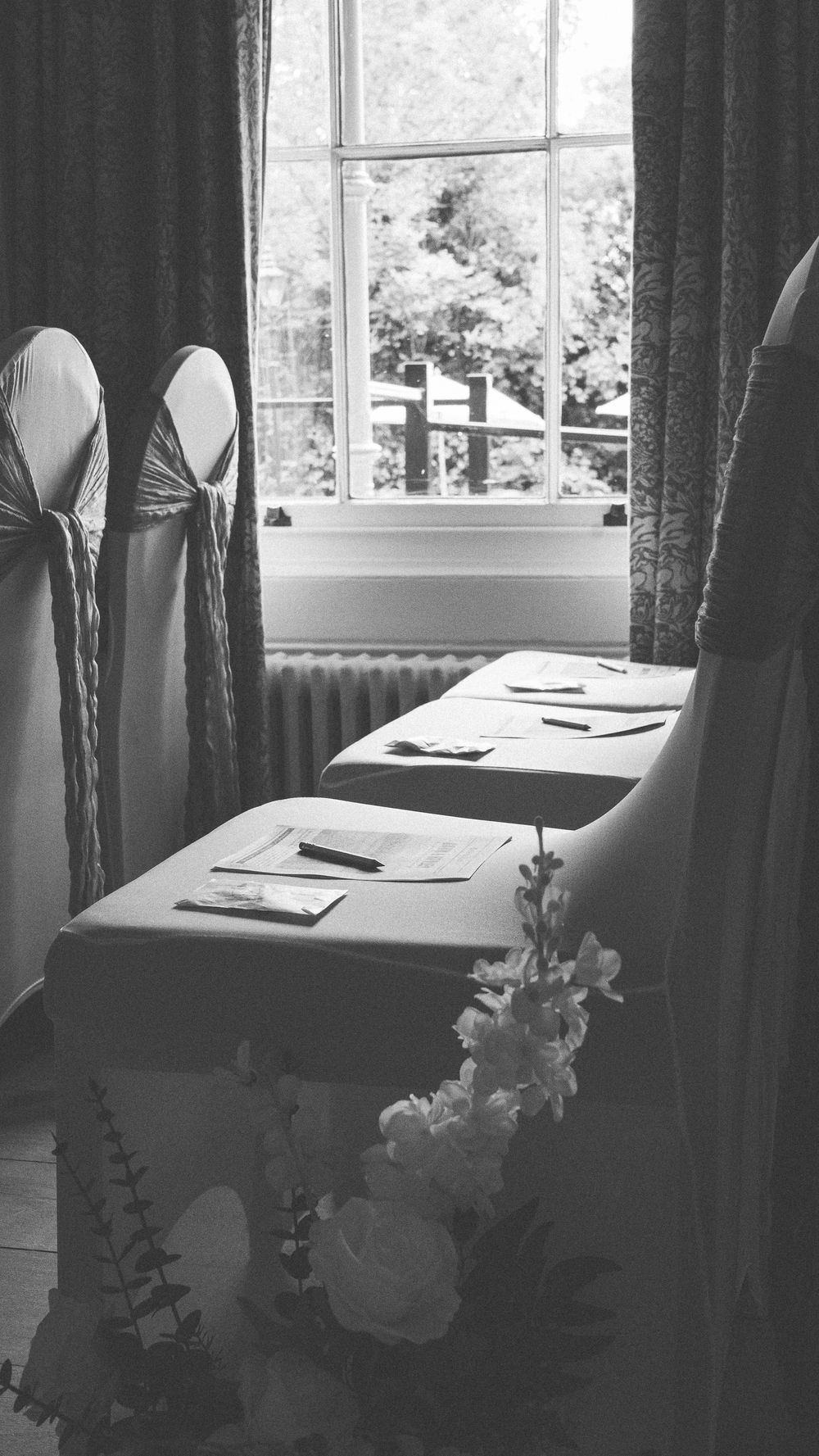 Elegant room setup with dark curtains and white tablecloths near bright windows with delicate floral arrangements.