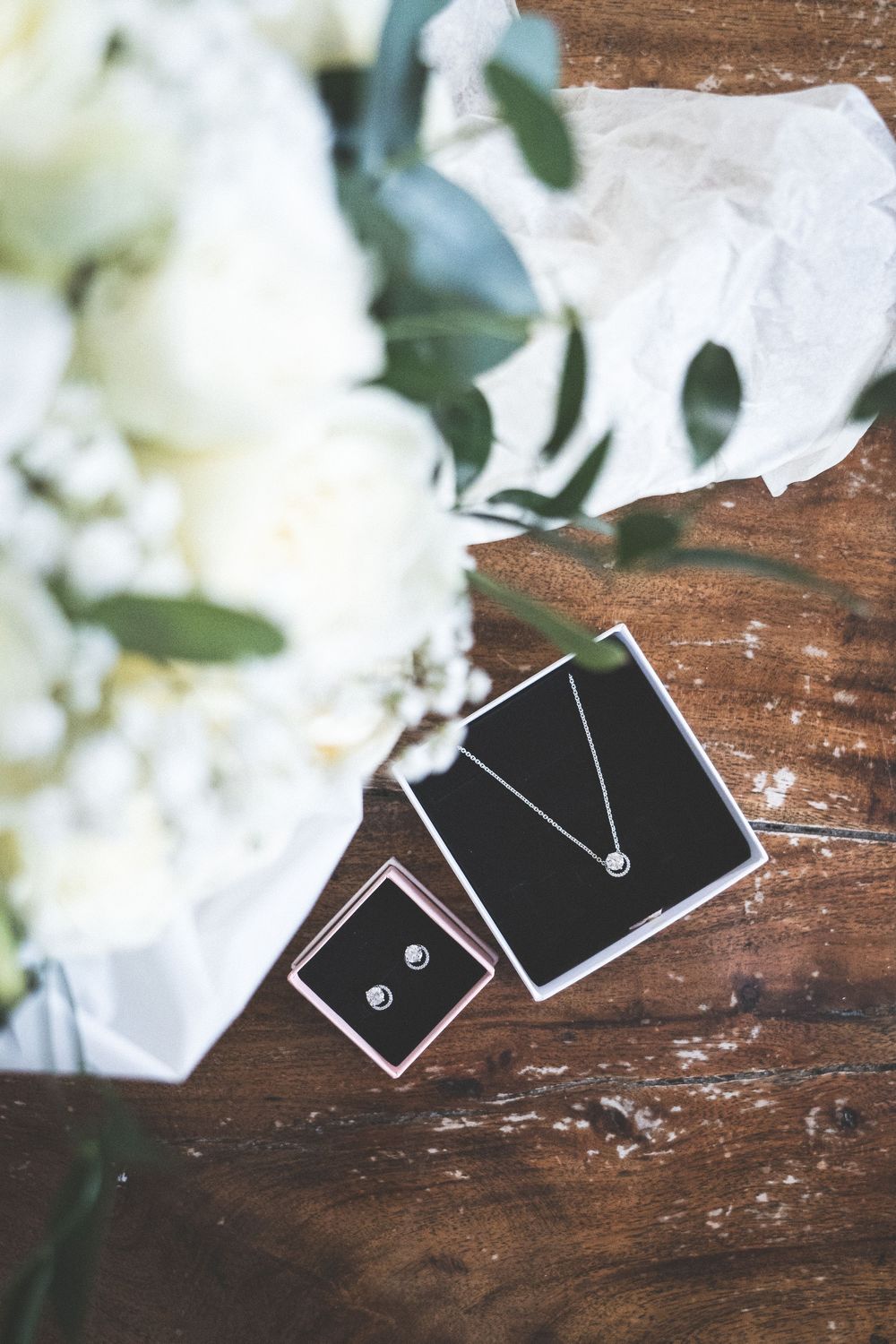 Silver necklace and earring gift boxes displayed with white flowers and greenery on rustic wooden surface.