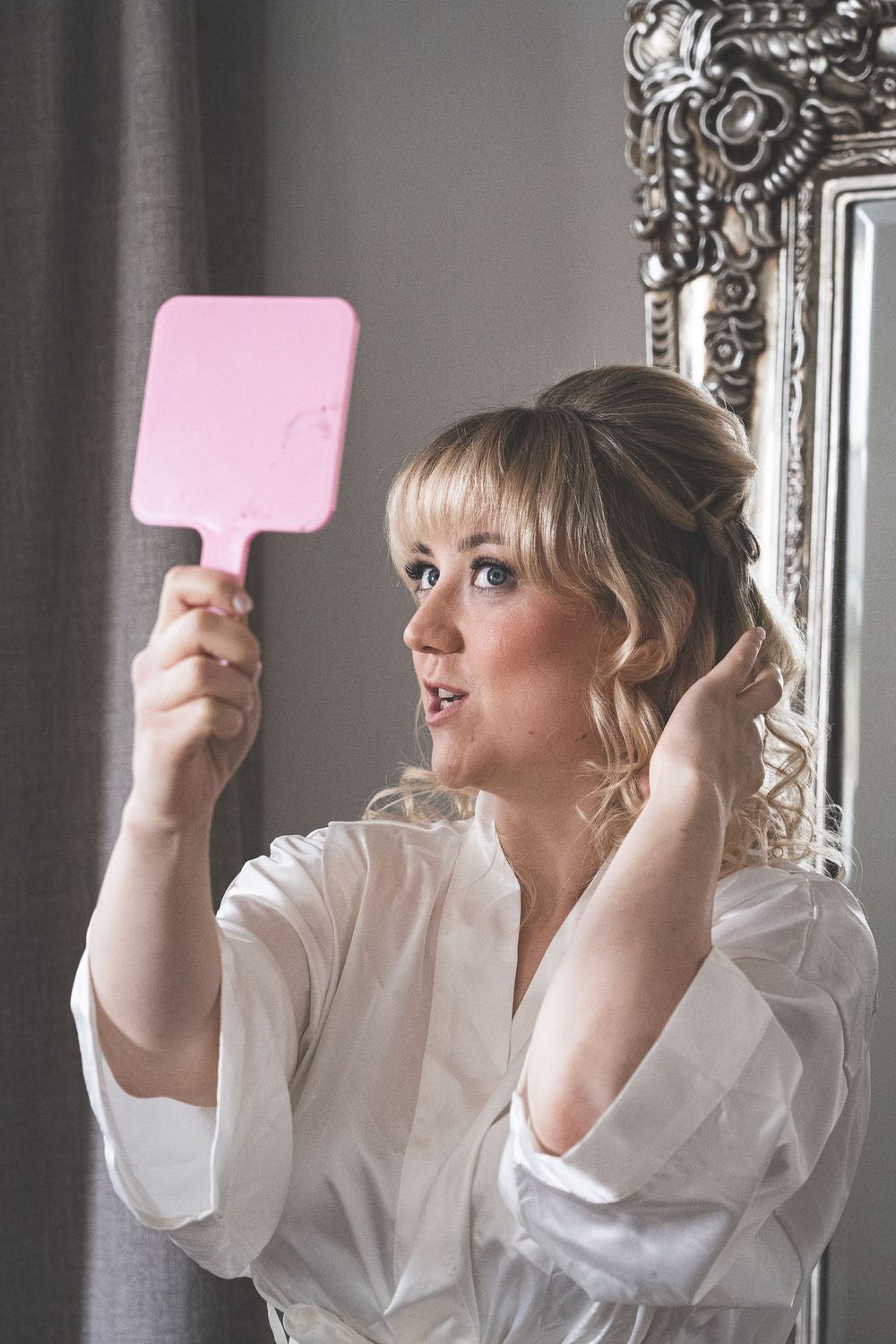 Someone holds up a pink handheld mirror while wearing a white robe and styling their hair in front of an ornate framed mirror.
