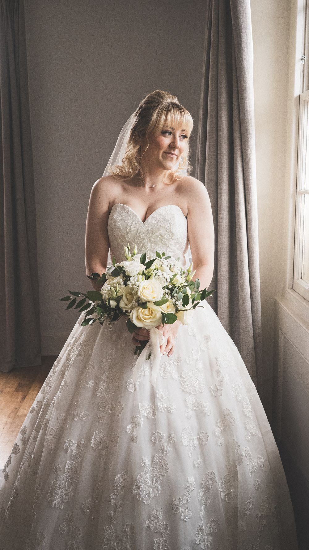 A bride in a stunning lace ball gown wedding dress holds a white rose bouquet near a sunlit window.