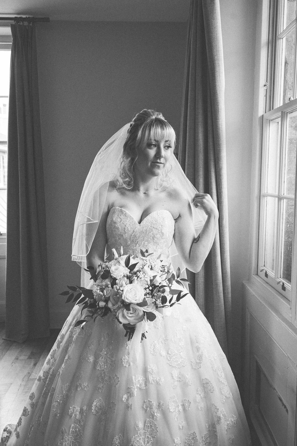 Black and white photograph of a bride in a sweetheart strapless lace gown holding a bouquet near a window.