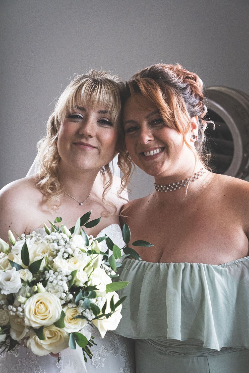 Two individuals in mint green dresses hold white rose bouquets while smiling together against a gray background.