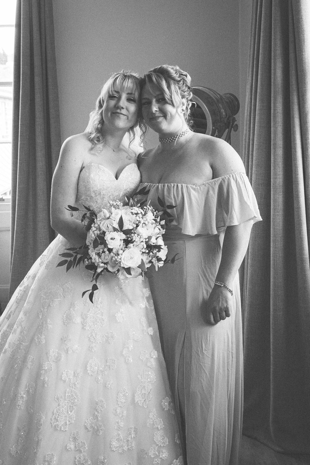 Black and white photo of bride in lace gown and bridesmaid in flowy dress sharing tender moment with bridal bouquet.