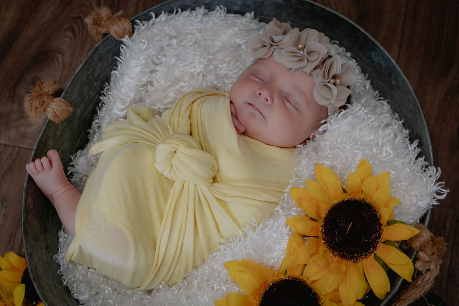 A sleeping newborn in a yellow wrap lies in a white furry basket with sunflowers and autumn leaves as decorations.
