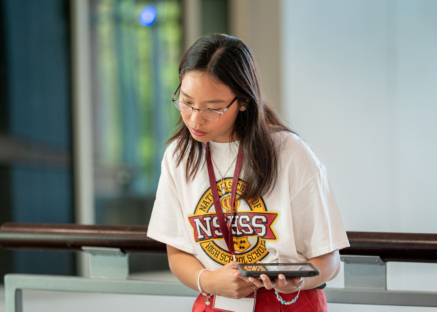 Student wearing NSEEG t-shirt looks down at digital device in an educational setting.