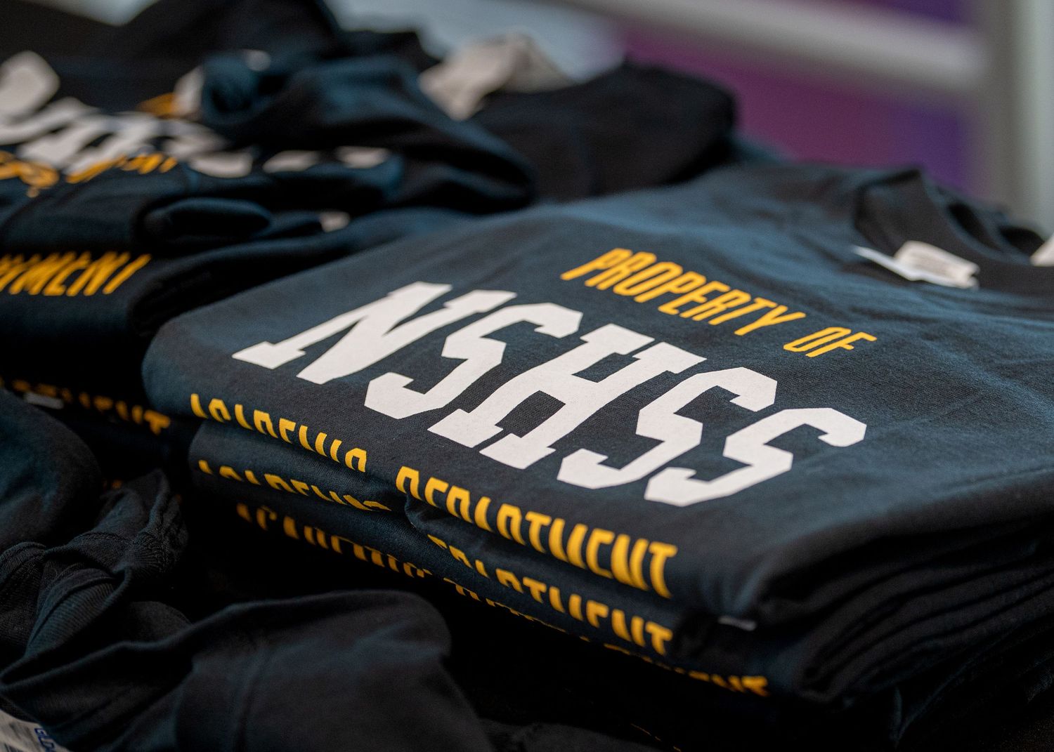 Stack of folded black NSEEG t-shirts with yellow text displayed for sale.