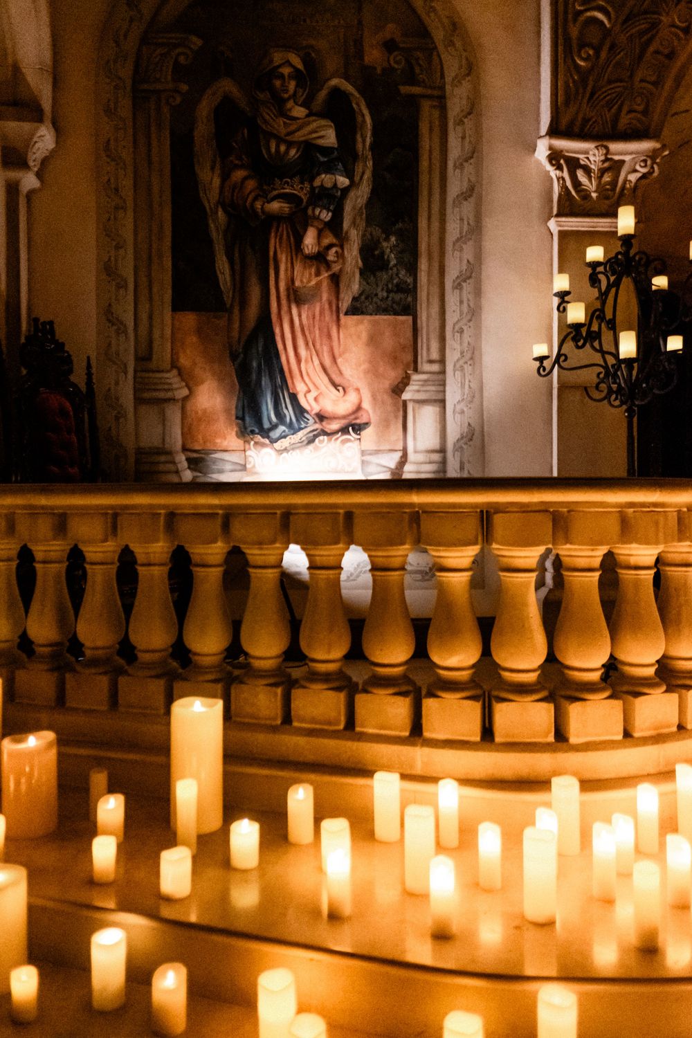 Candlelit church altar with angel mural and rows of glowing white pillar candles.