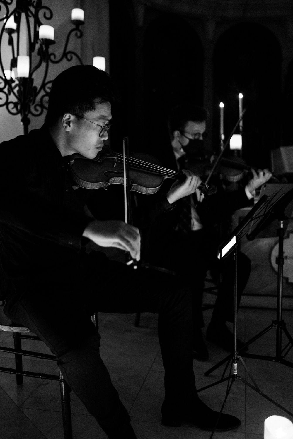 Black and white photo of two violinists performing by candlelight in a church.