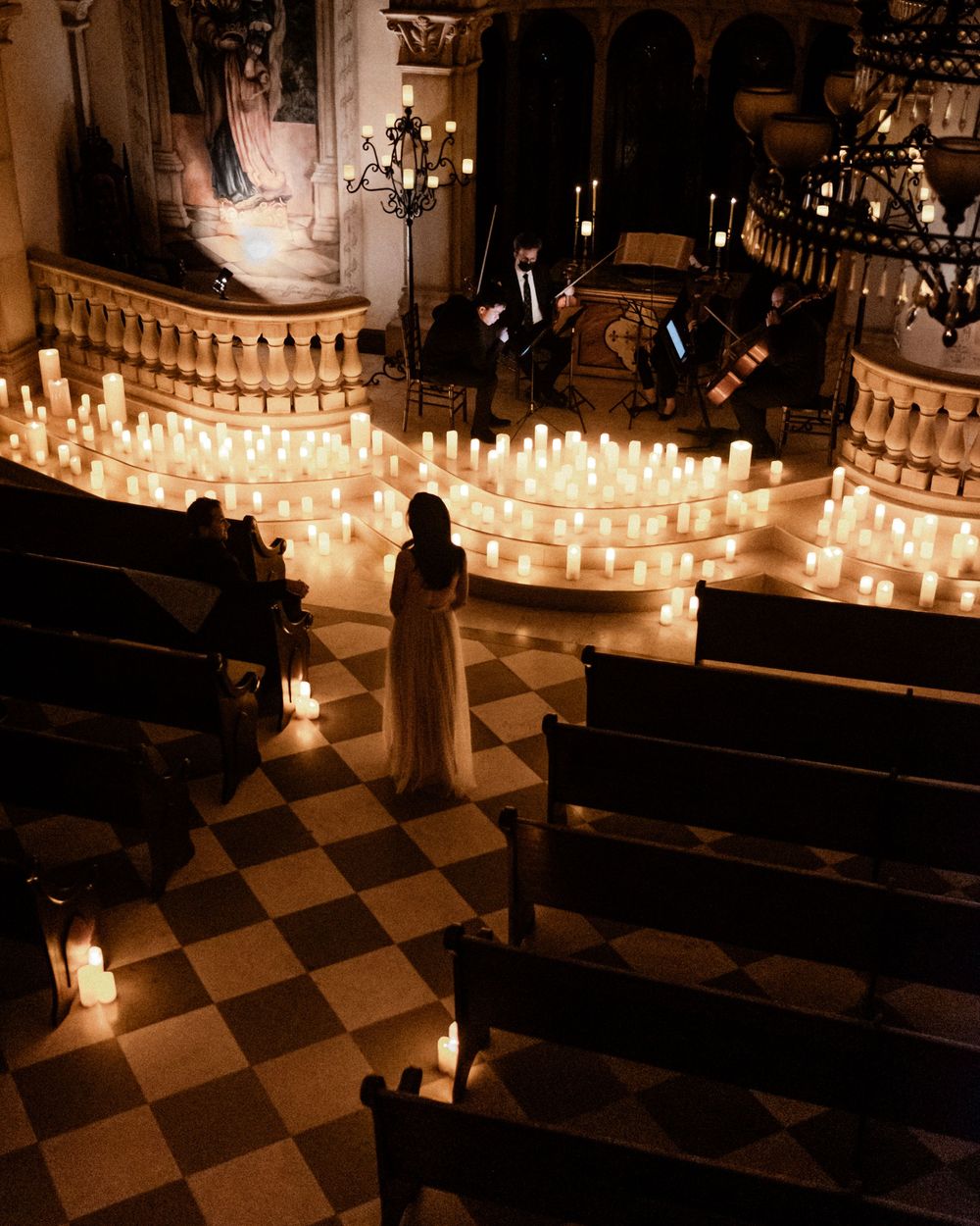 Bride standing in candlelit church aisle surrounded by glowing candles and string quartet.