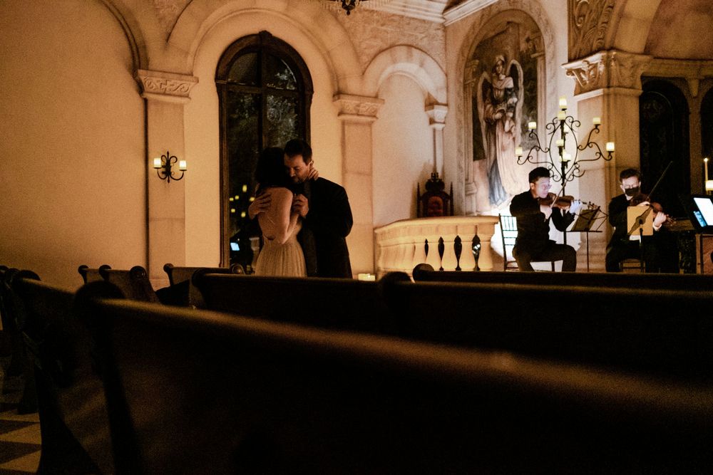Couple sharing an intimate kiss in a dimly lit historic church with a violinist performing in background.
