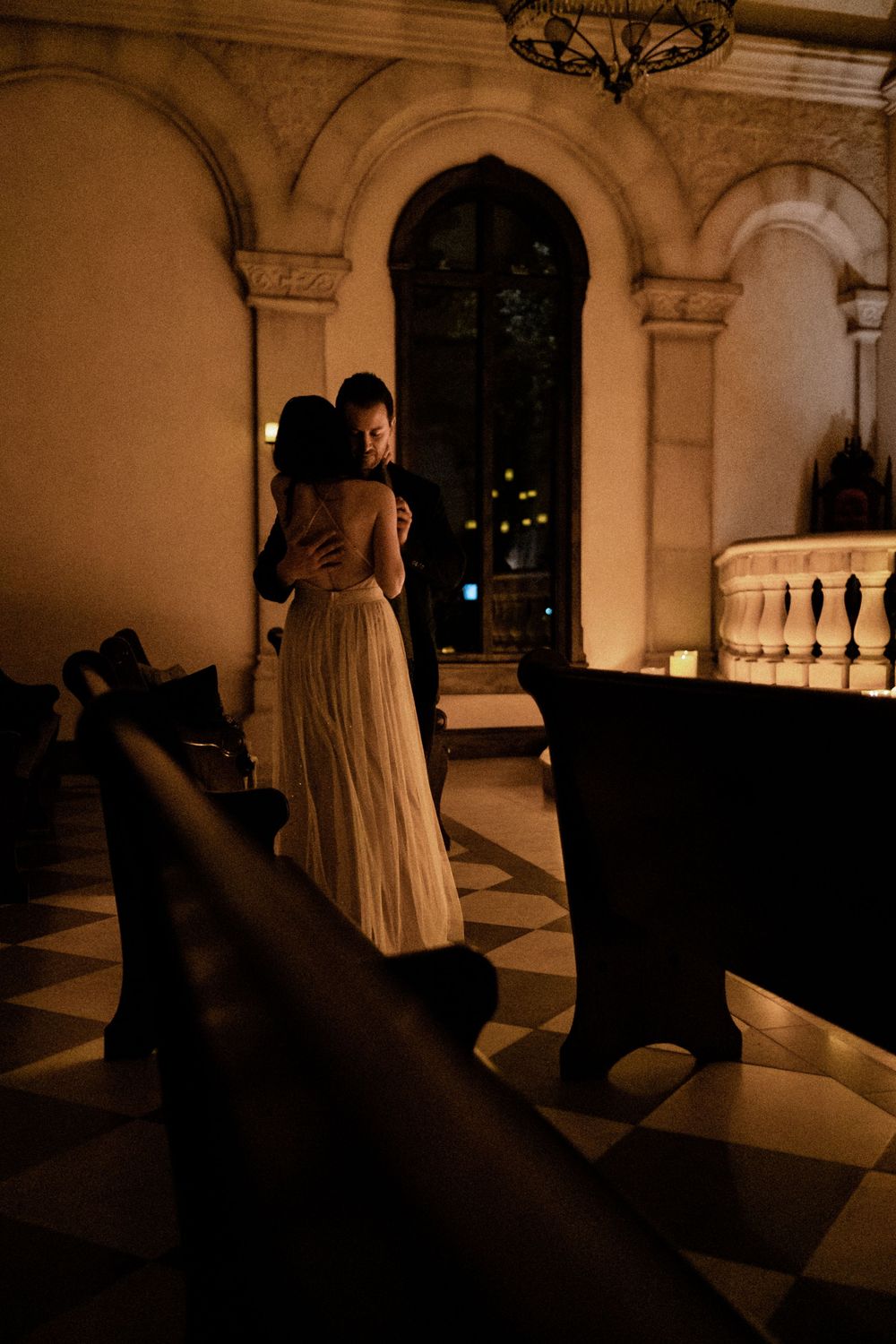 Couple embracing in a candlelit ornate church with checkered marble floors and arched stone windows.