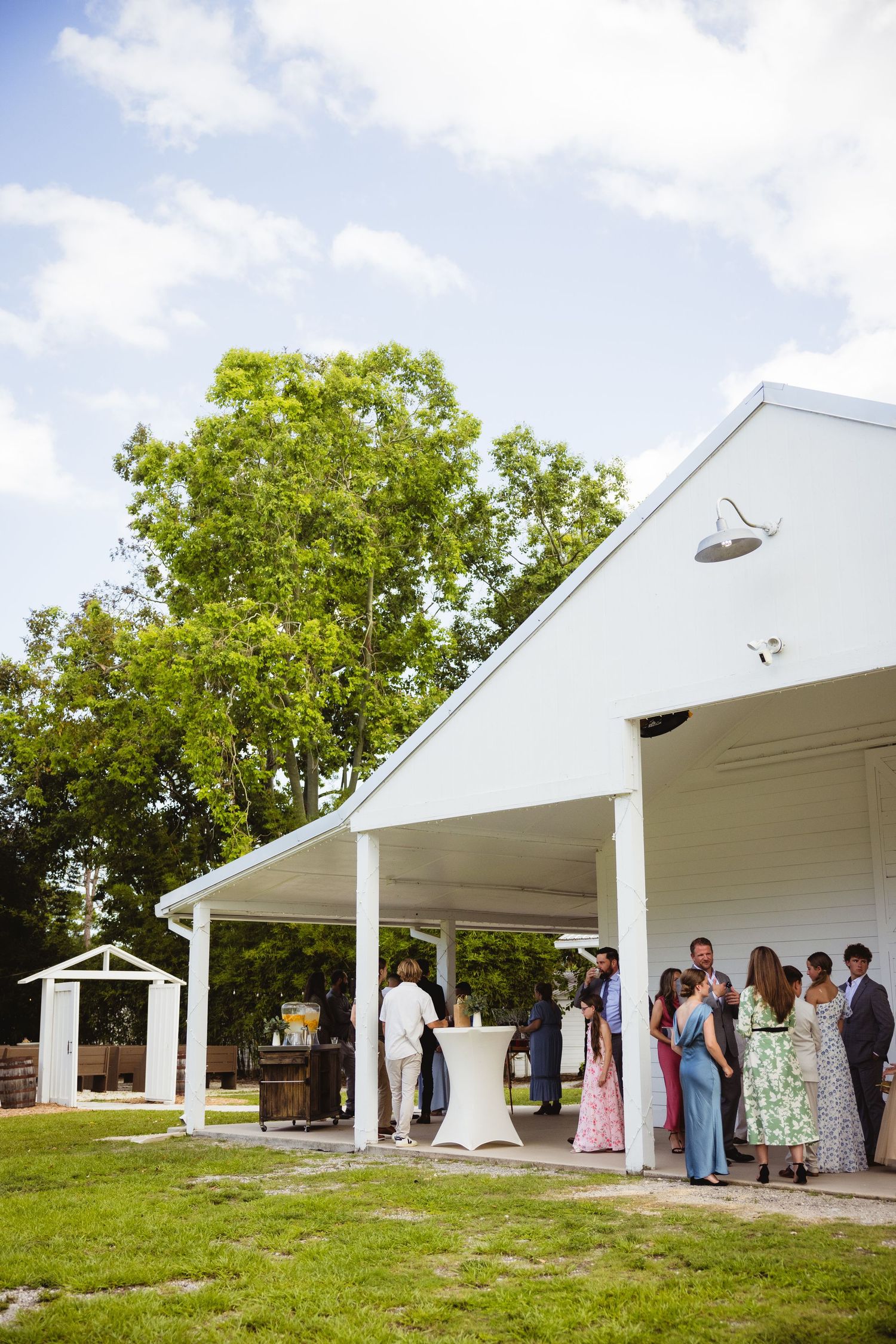 Wedding guests gather under a white covered porch next to a green lawn and tall trees on a sunny day.