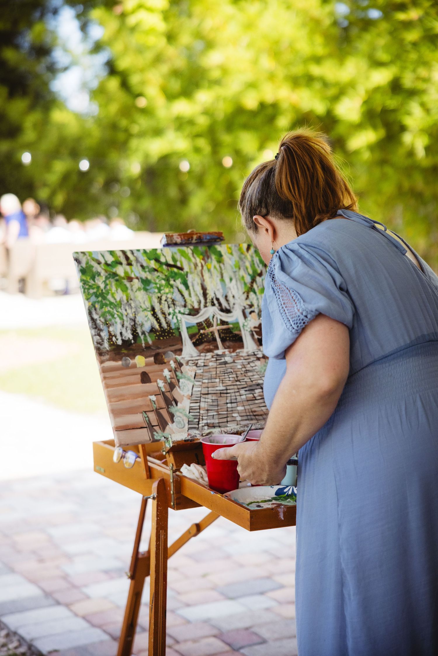 An artist in a blue dress paints at an outdoor easel while working on a detailed architectural scene in a sunlit park.