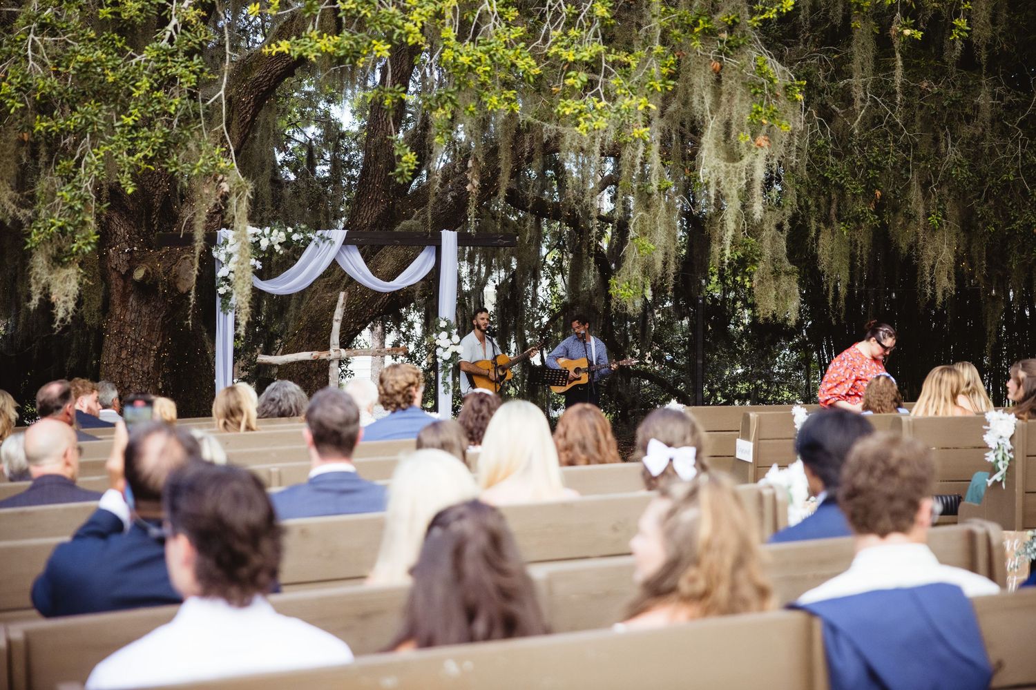 A panoramic view shows an outdoor ceremony beneath Spanish moss-draped trees at a garden venue.