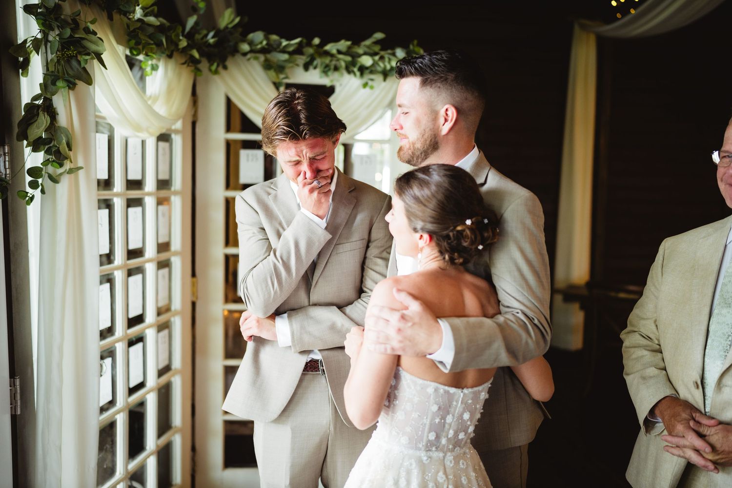 An emotional moment between two figures in formalwear underneath an archway decorated with greenery at a wedding ceremony.