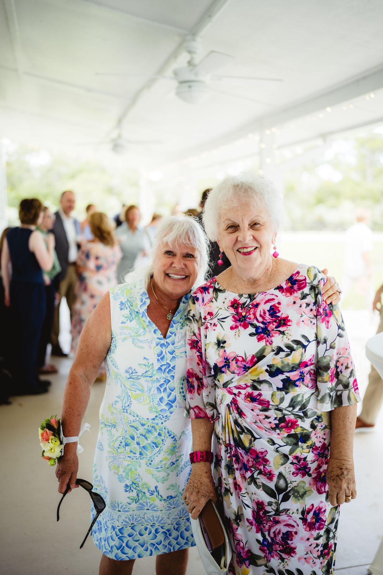 Two elegantly dressed ladies in floral print dresses smile together at a social gathering under a covered venue.