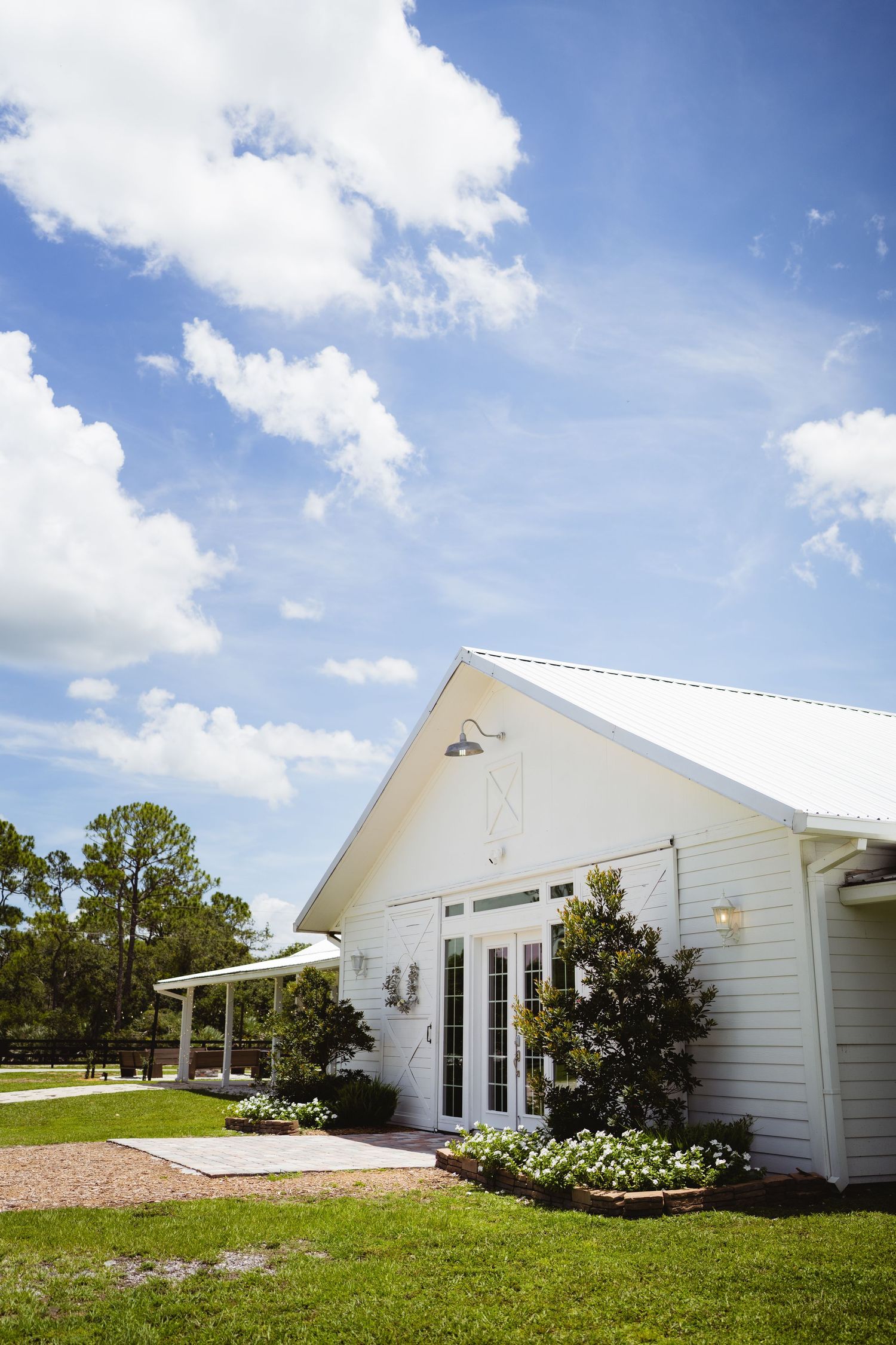 Charming white farmhouse venue with French doors, covered porch and manicured lawn under bright blue skies with puffy clouds.
