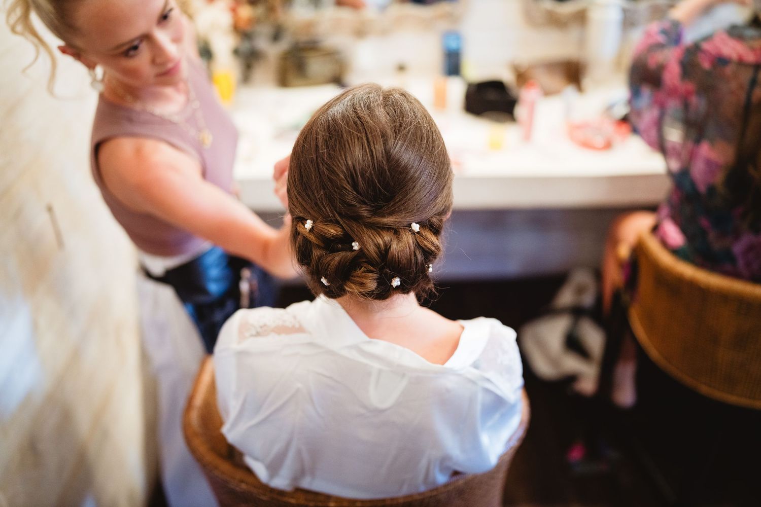 A bride prepares for her wedding while a hairstylist styles her updo in a bright bridal suite.