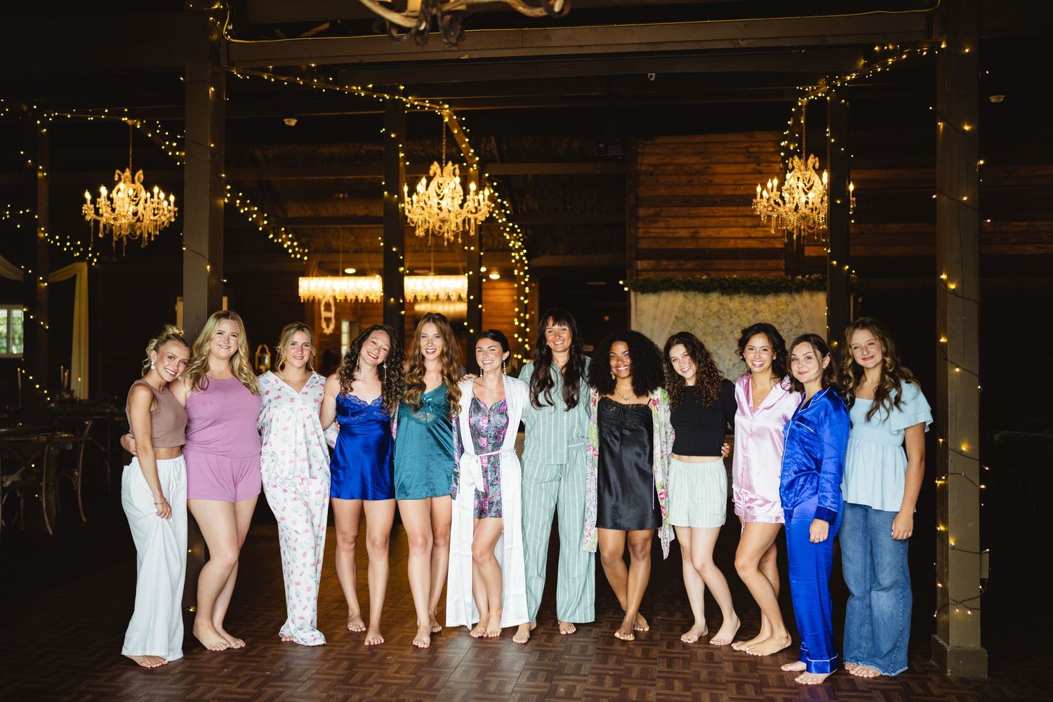 A group of people pose together in a rustic indoor venue with hanging chandeliers and decorative string lights.