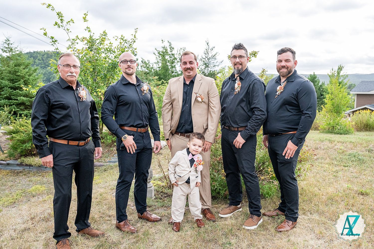 Groomsmen in dark suits pose together with a small child in formal attire against an outdoor natural background.