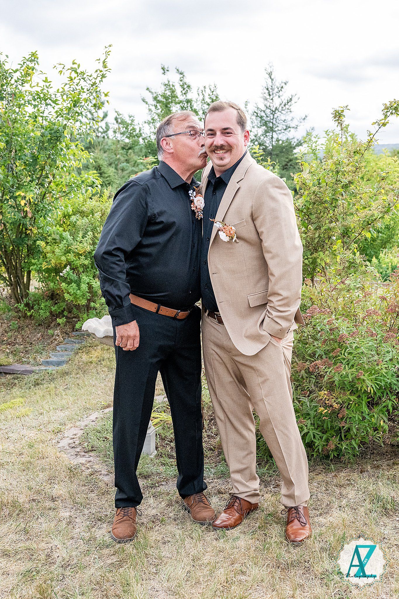 Two people in contrasting outfits, black and tan, share a light-hearted moment together in an outdoor garden setting.