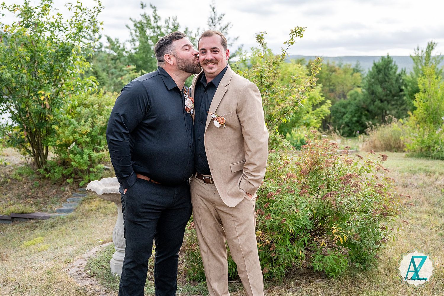 Two people share a moment in casual and formal attire while standing in a scenic outdoor garden setting.