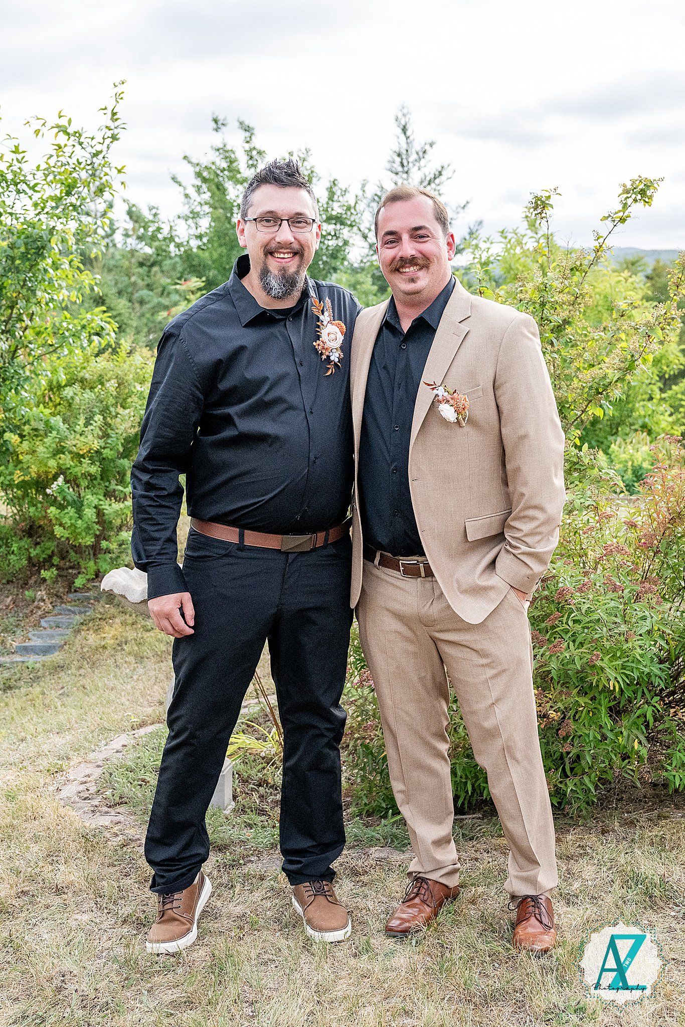 Two men in formal attire with matching floral boutonnieres pose outdoors against greenery.