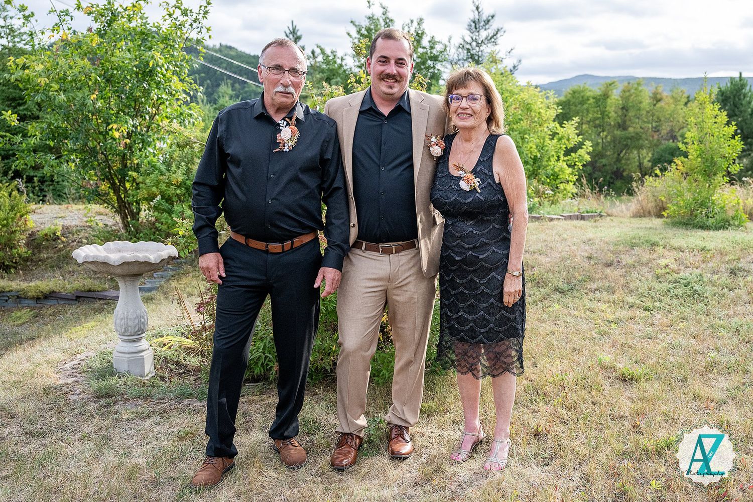 Two men and a woman pose for a photo outdoors in a garden setting with lush green foliage in the background.