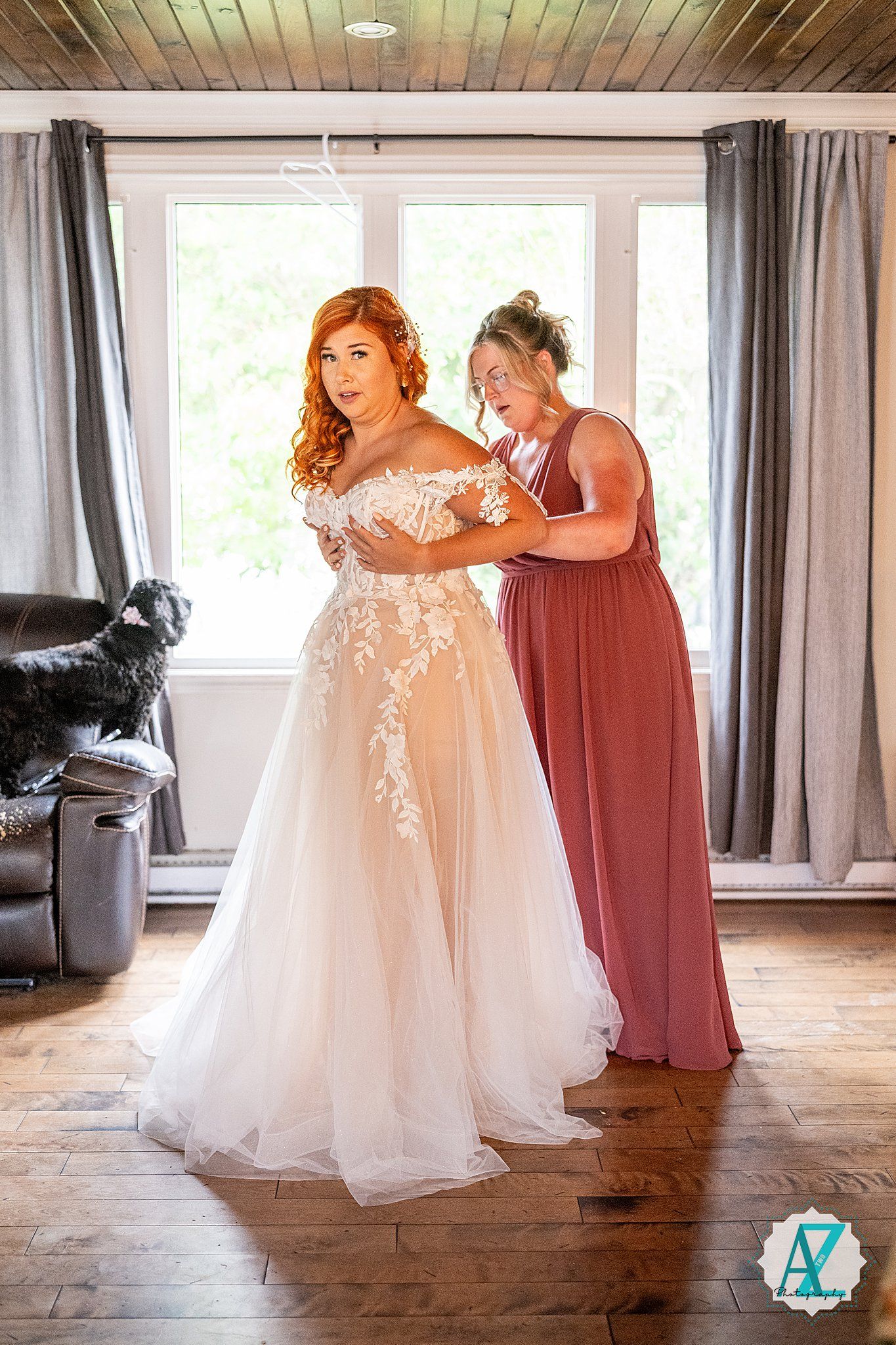 Wedding preparation scene showing a long white bridal gown being adjusted in a rustic room with wooden floors and windows.