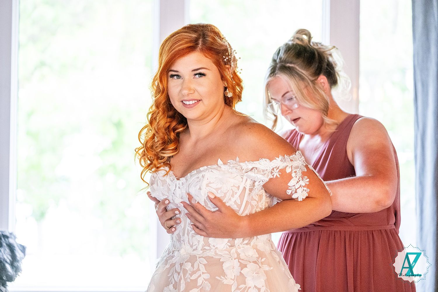 Wedding party members in dusky rose gowns assist with arranging a bride's gown in natural lighting.