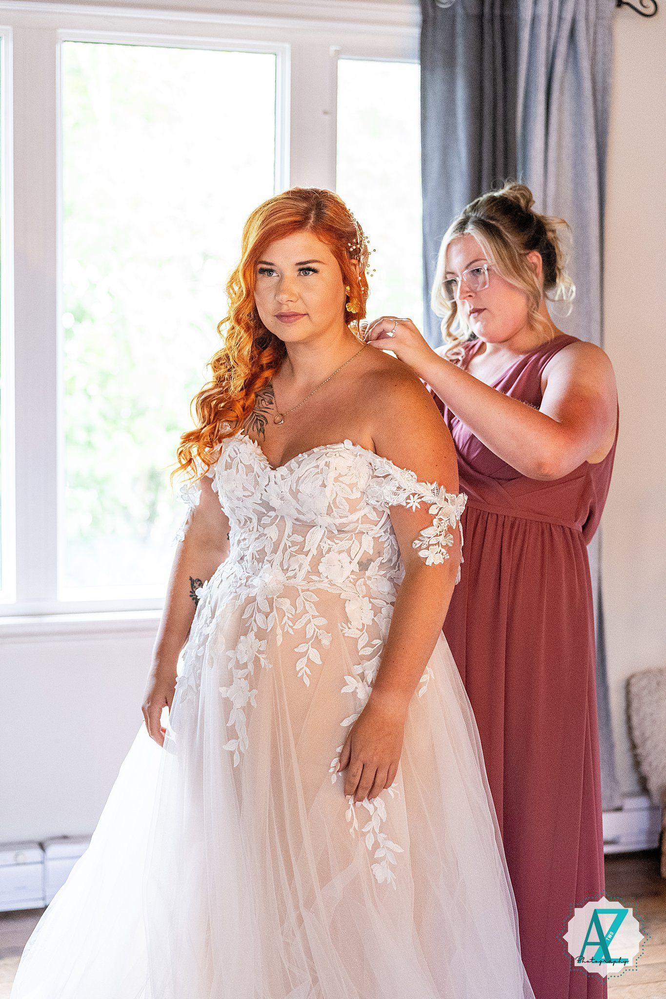 The bride gets ready in a beautiful lace wedding gown while assisted by her bridesmaid in dusty rose.