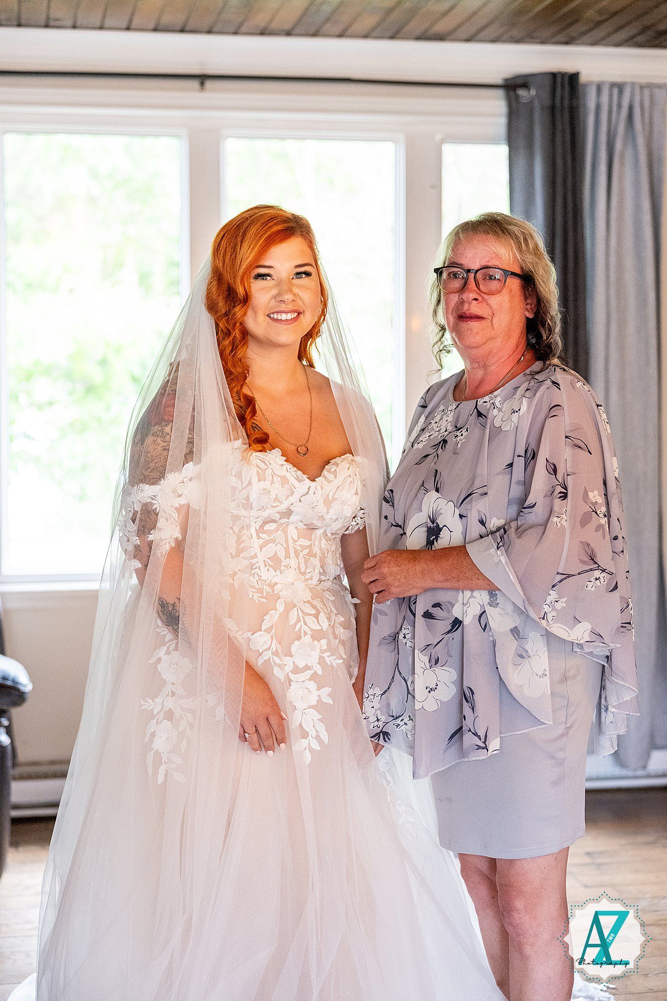 A bride in a stunning lace wedding gown poses next to someone wearing a floral patterned dress in a bright sunlit room.