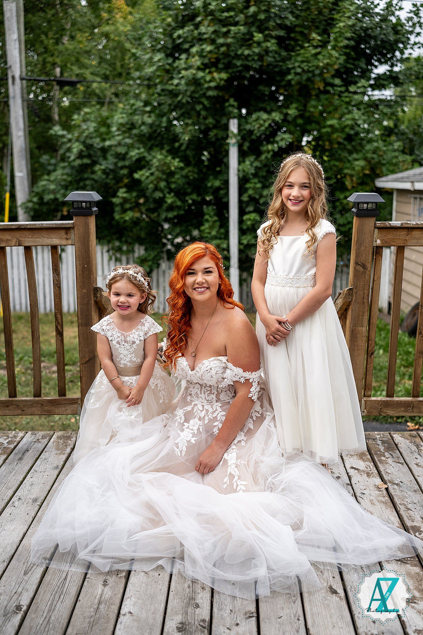 A group poses together on a wooden deck surrounded by lush greenery wearing coordinated white dresses.