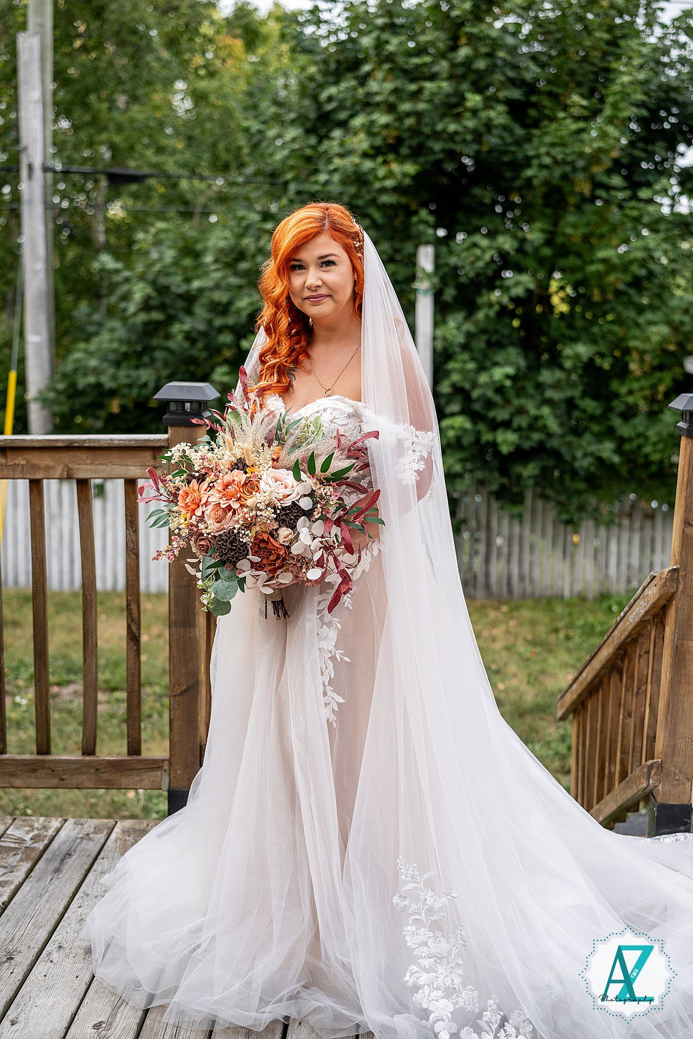 A bride in a white flowing gown and veil sits on a wooden deck surrounded by lush greenery holding an autumn bouquet.