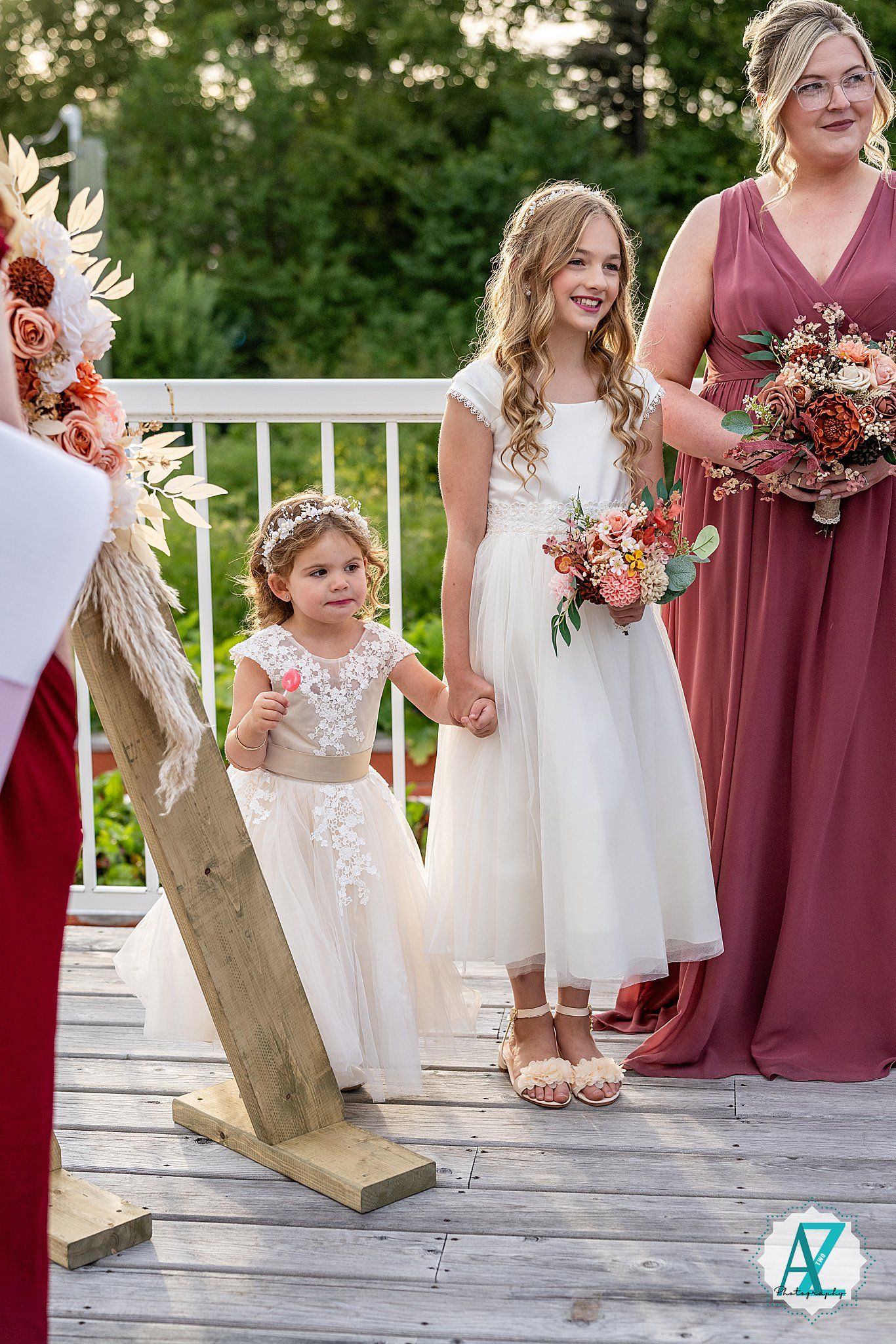 Young flower girls in white dresses and bridesmaids in mauve gowns standing together at outdoor wedding ceremony.