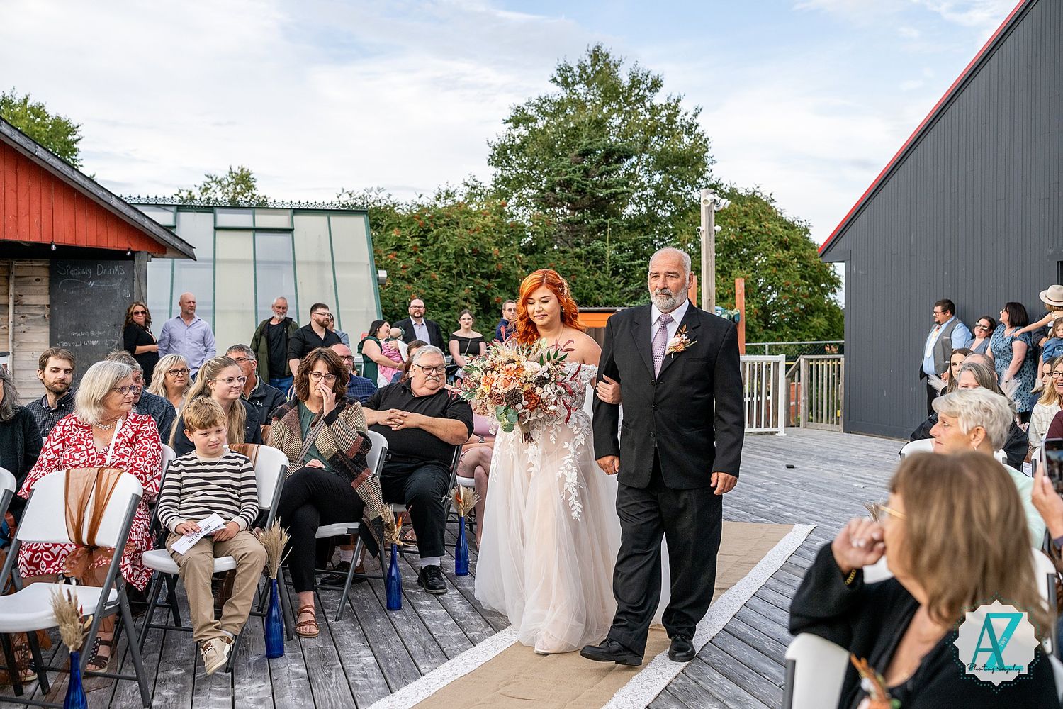 Wedding party stands in front of modern gray and red barn during outdoor celebration.