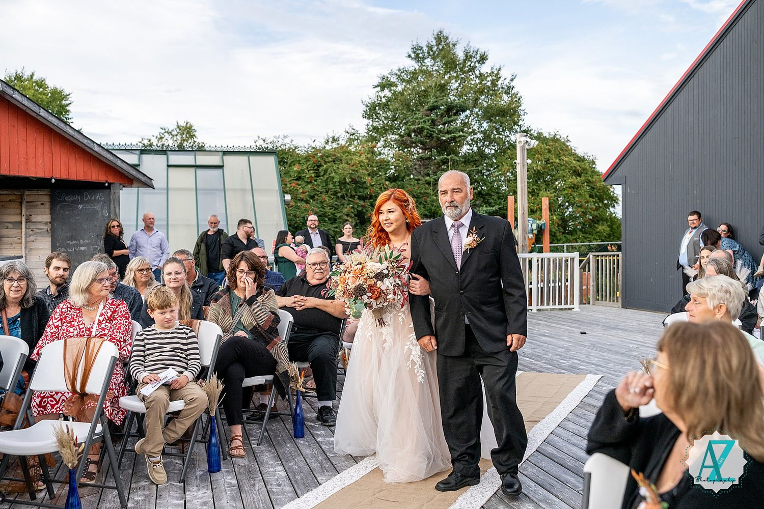 Wedding ceremony guests seated outdoors at a rustic venue with red and grey buildings in the background.