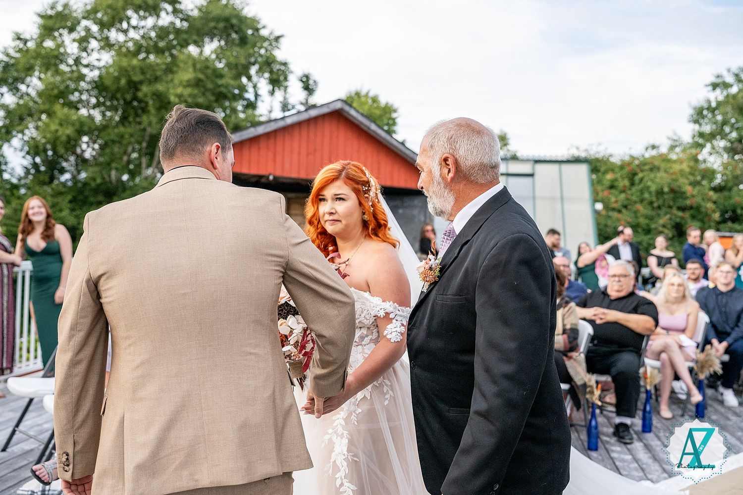 Outdoor wedding ceremony taking place in front of a red barn with guests seated on white chairs.