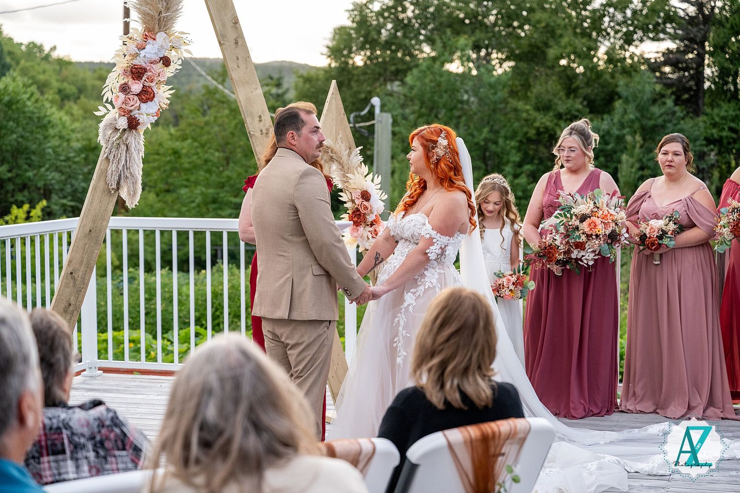 Wedding party poses outdoors with bridesmaids in burgundy dresses and tan suits on a sunny day.
