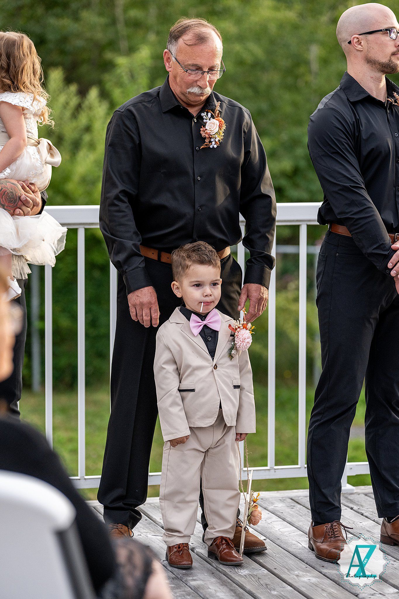 A young child in a beige suit stands with adults on a wooden deck during a formal event.