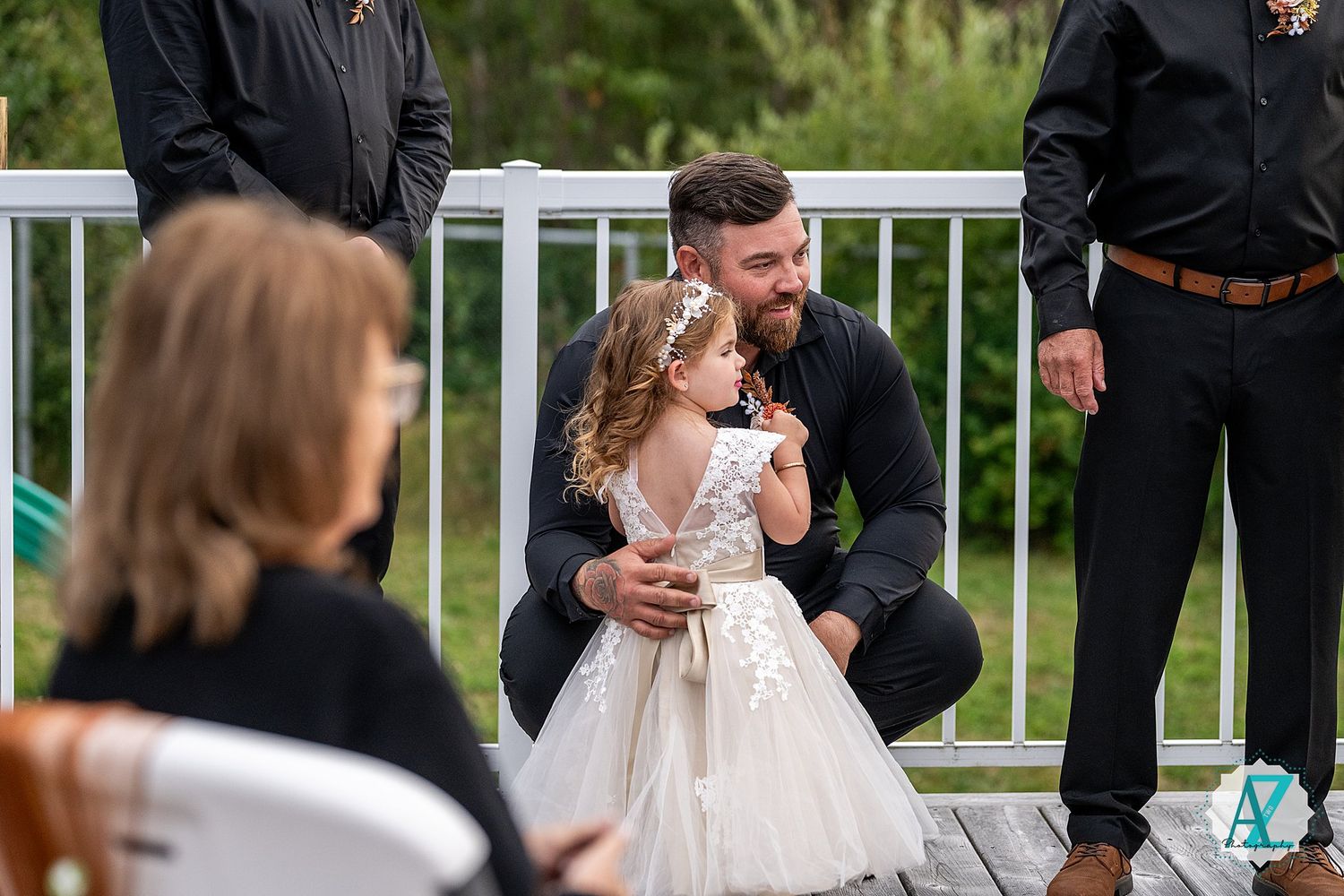 Outdoor wedding ceremony with guests seated on white chairs on a wooden deck surrounded by greenery.