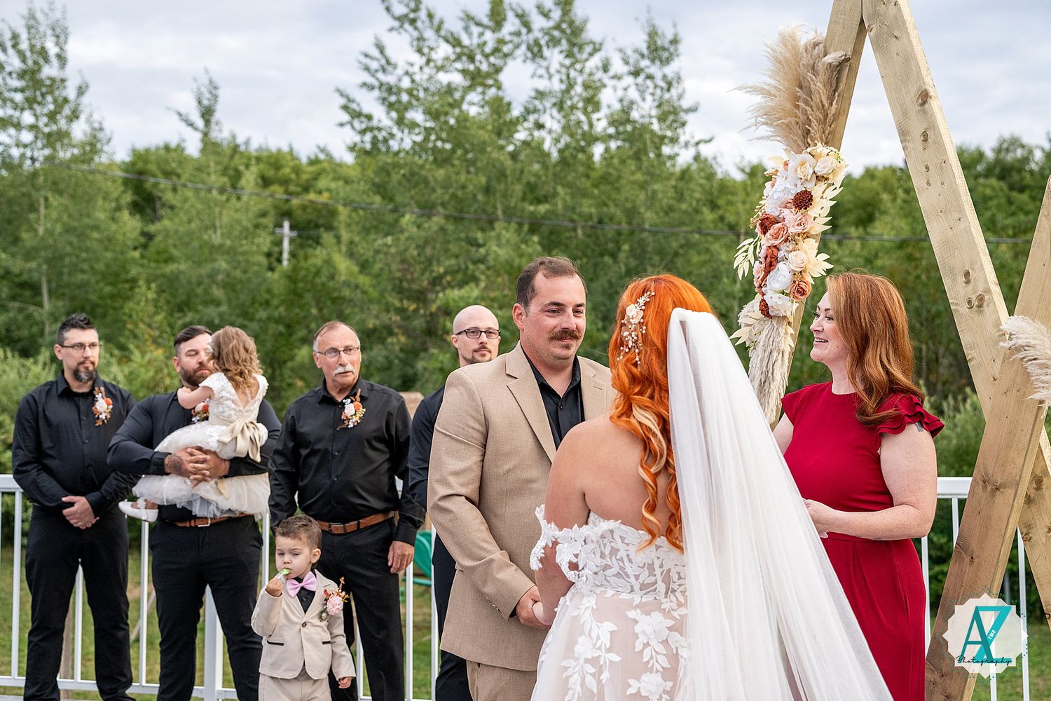 Wedding ceremony guests gathered outdoors under blue sky with wooden arch and floral decorations.