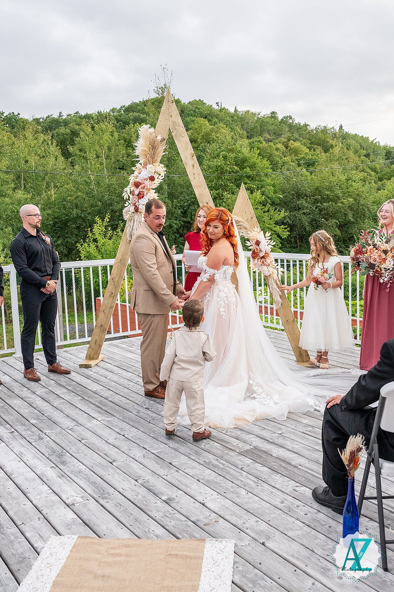 A wedding ceremony unfolds on a white wooden deck overlooking lush green hills on a cloudy day.