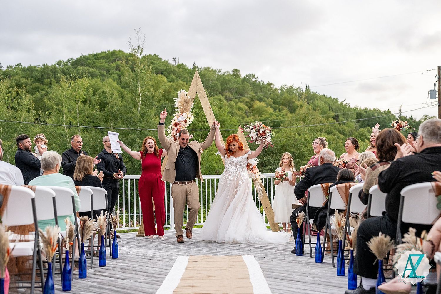 Wedding celebration on a mountain deck with guests cheering as couple celebrates under wooden arch.