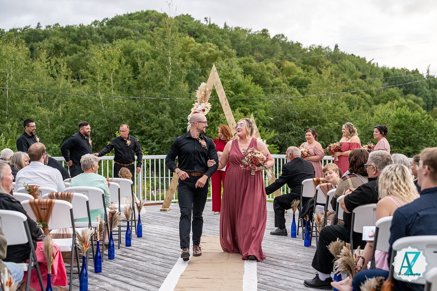 Wedding processional on an outdoor deck surrounded by green mountains and guests in attendance.
